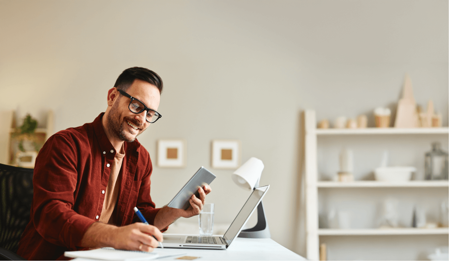 "Homem sorridente vestindo camisa casual, trabalhando em uma mesa com laptop e tablet, em um ambiente doméstico organizado."